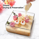 A clear square glass container filled with sand or powder, shown with preserved flowers arranged inside. A hand pours more powder into the container from a small glass cup.