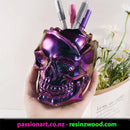 Purple skull-shaped pen holder with pens held by a hand, against a light background with flowers.